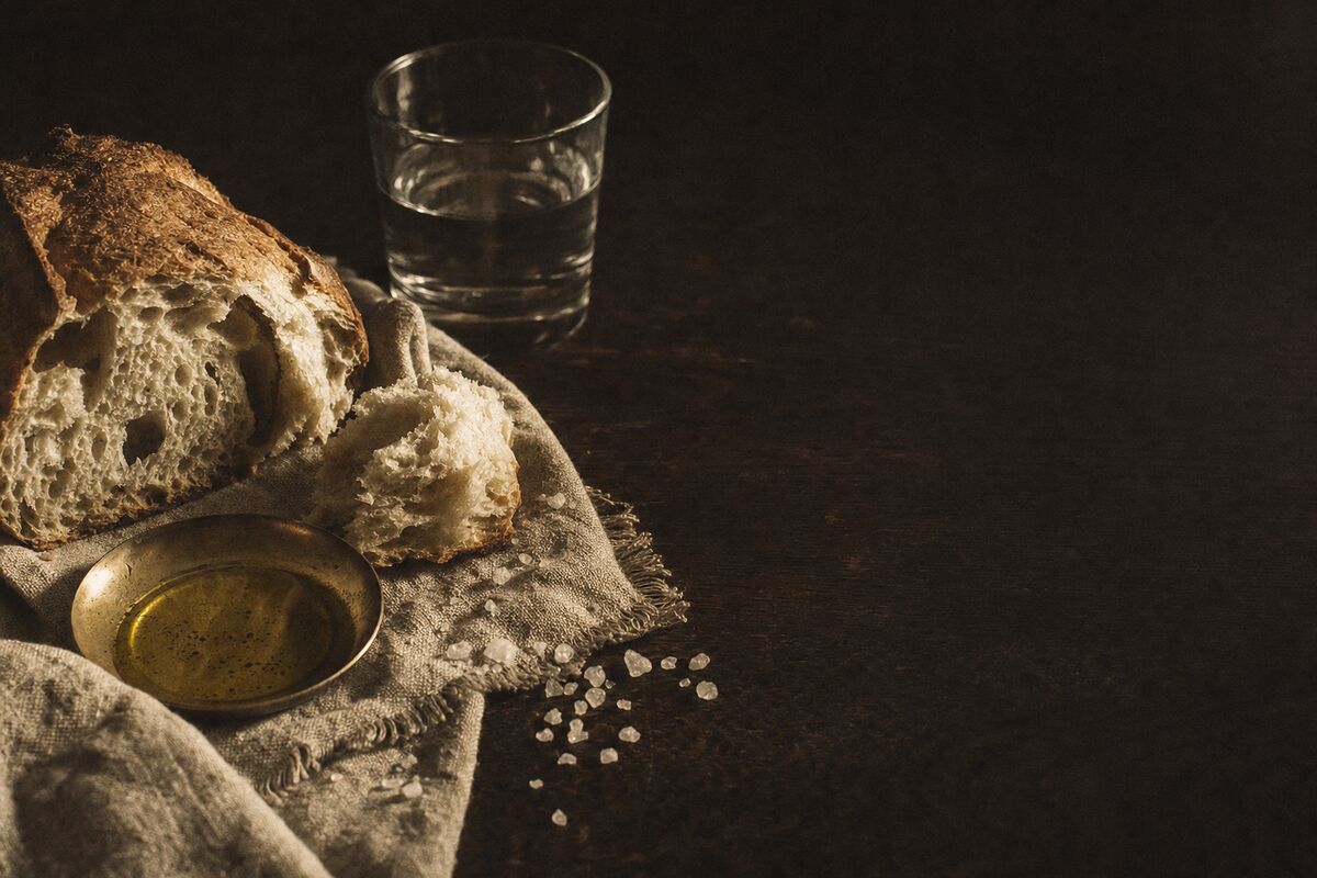 Torn country bread, a brass dish of olive oil and coarse salt on rough linen, lit from one side against a dark oak table.